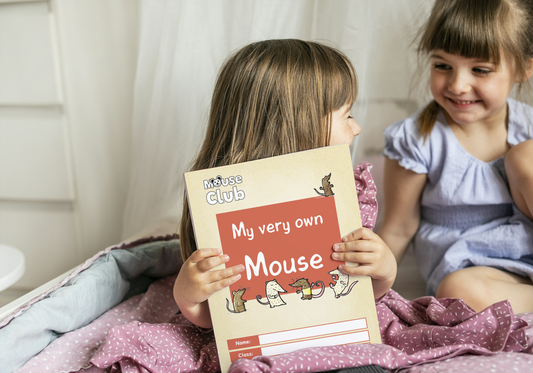 Two young girls are smiling at each other while one holds out a scrapbook with the title "My very own Mouse". 