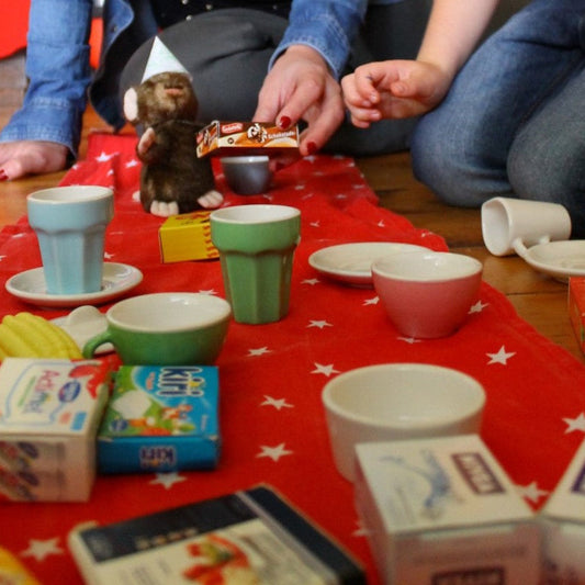 A parent and child are playing picnic with their toy mouse. Mouse is wearing a party hat. 