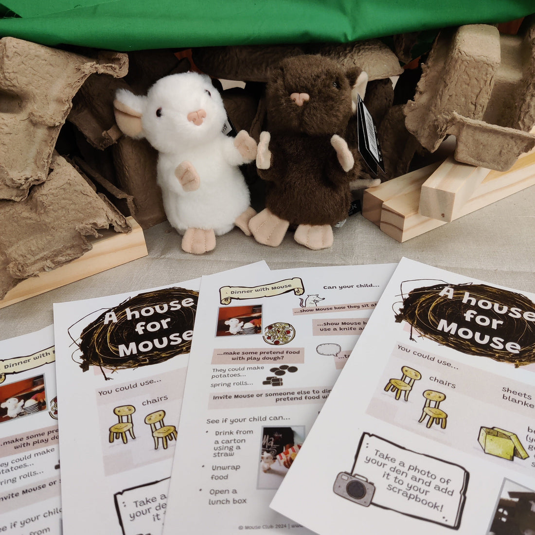 Toy Mouse sit in front of a cardboard house. Educational materials are on the table. 
