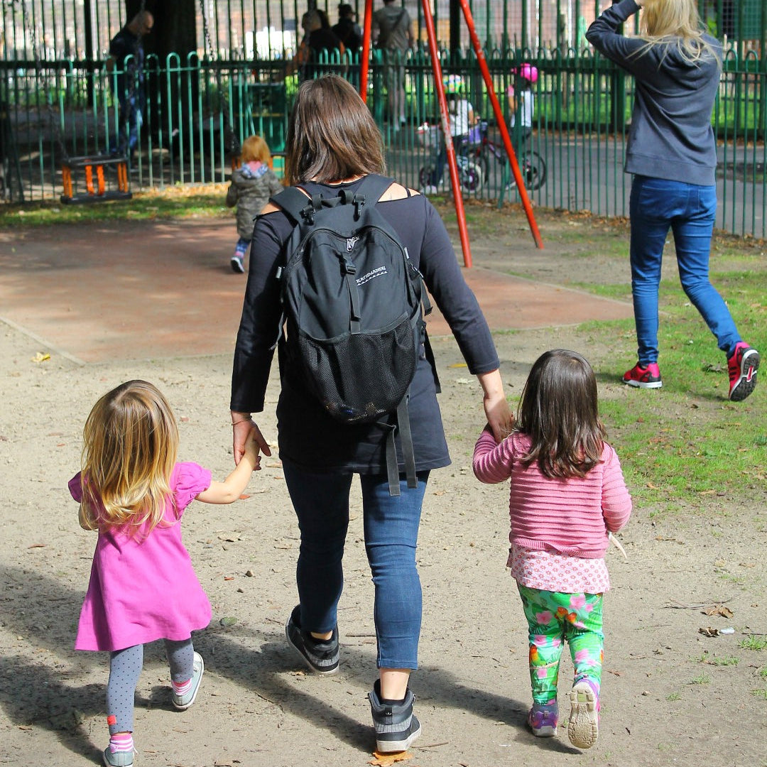 A mum leads her two daughters to the park. They are walking away from the camera and holding hands.