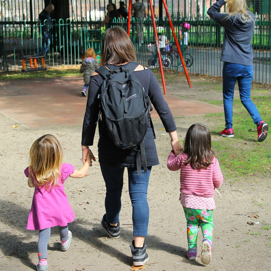 A mum leads her two daughters to the park. They are walking away from the camera and holding hands.